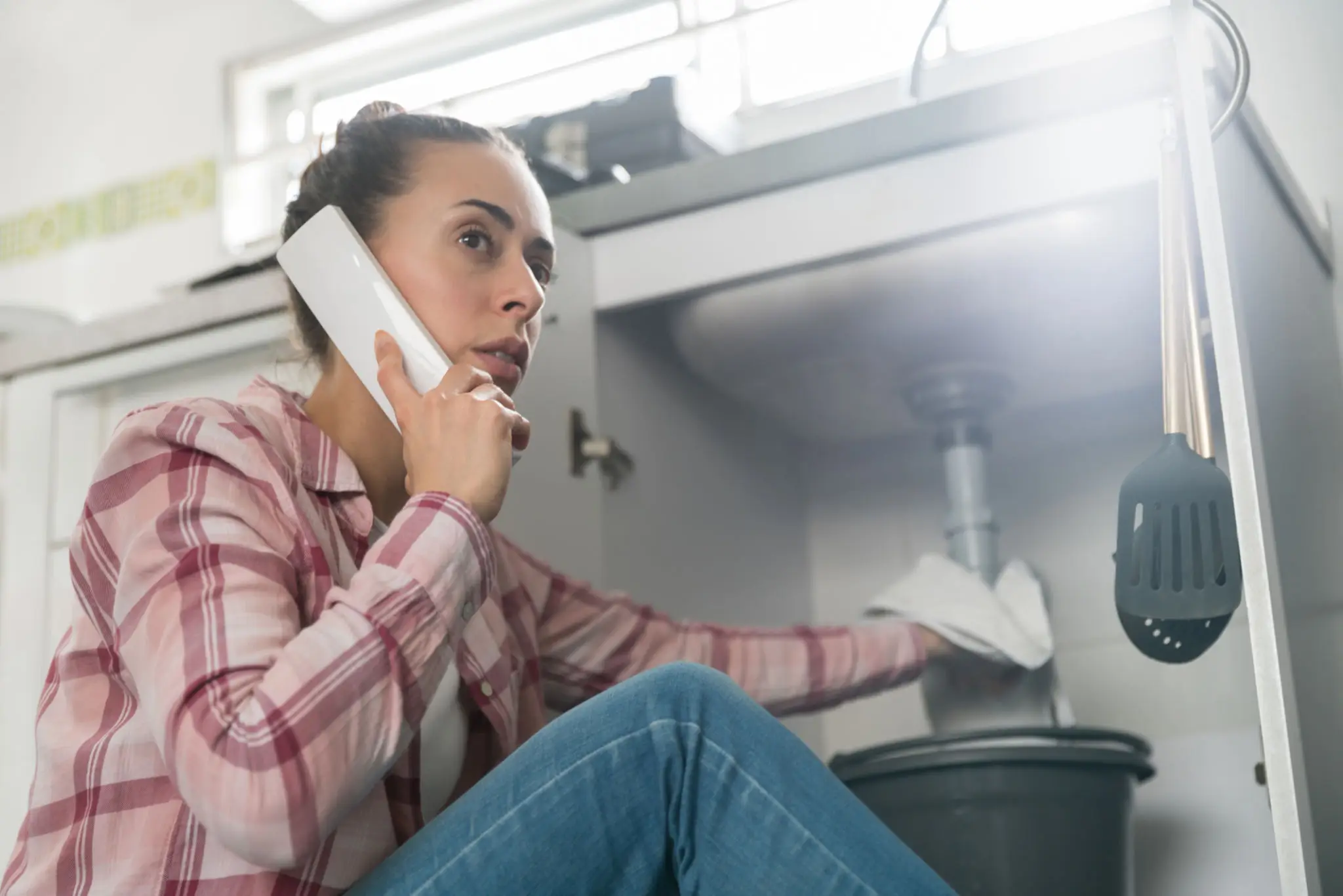 Woman on phone fixing kitchen sink plumbing